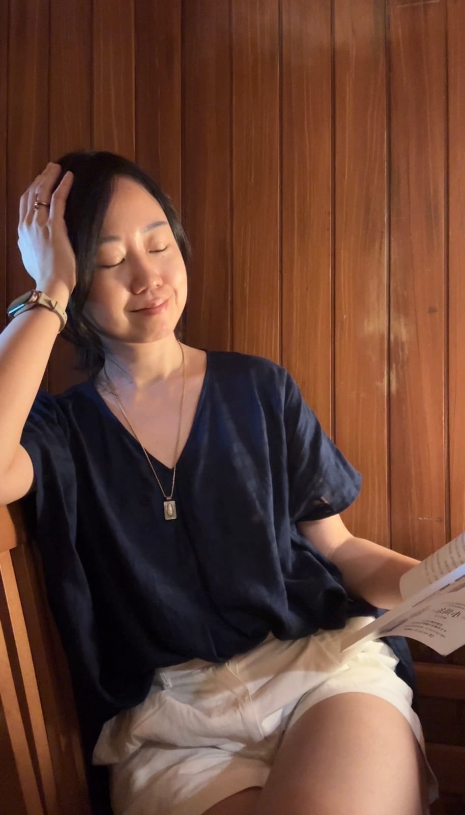在翫雙溪書院的木牆前坐著，閉眼撥著頭髮、手裡拿著一本書