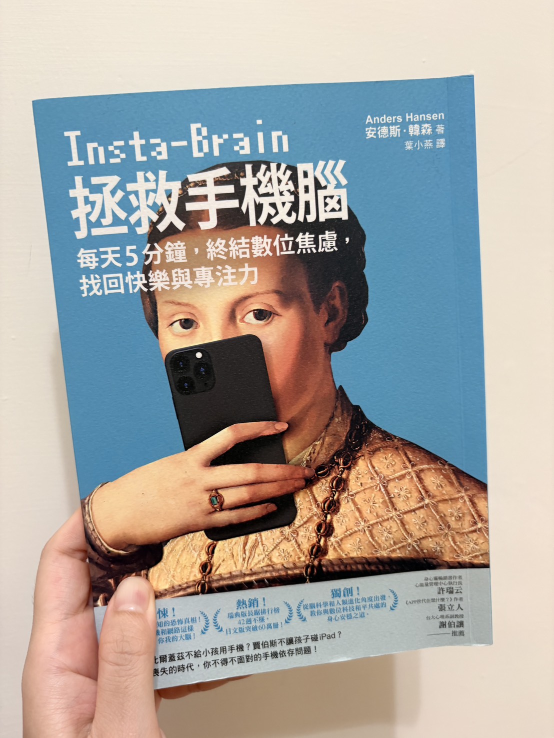手拿著《拯救手機腦》這本書，封面是文藝復興肖像畫人物舉著手機自拍